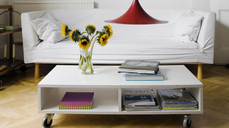 White coffee table on wheels, books and magazines inside, vase of sunflowers, white couch in background