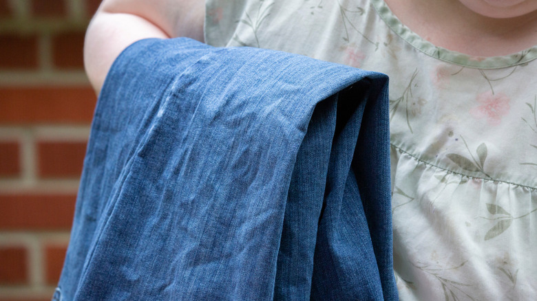 person wearing floral shirt holding up pair of wrinkled jeans against a brick background