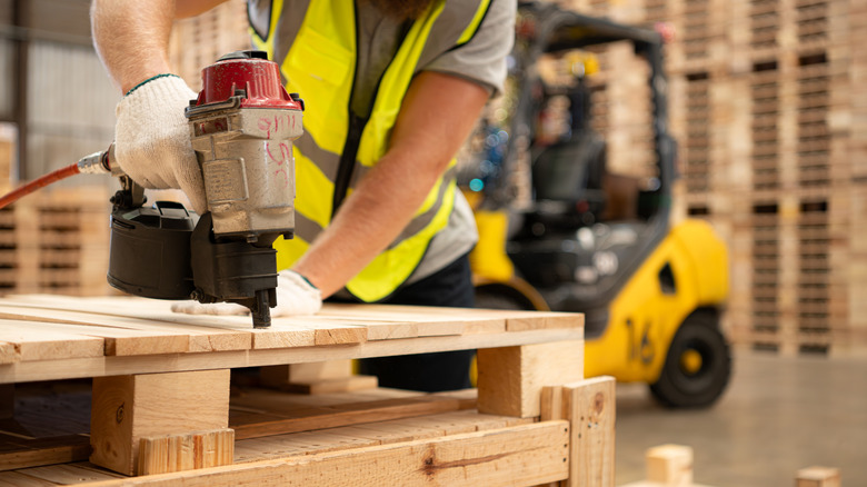 A construction worker in a high-visibility vest attaches nails into a wood pallet.