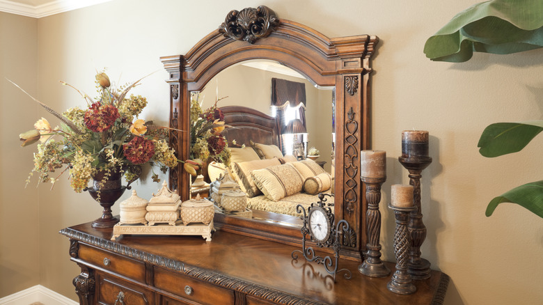 An ornately-carved wooden dresser with matching mirror sits in an outdated bedroom. The top is decorated with wooden candle holders, various knickknacks and a faux flower arrangrment. You can see some of the bed and window dressing through the mirror's reflection.