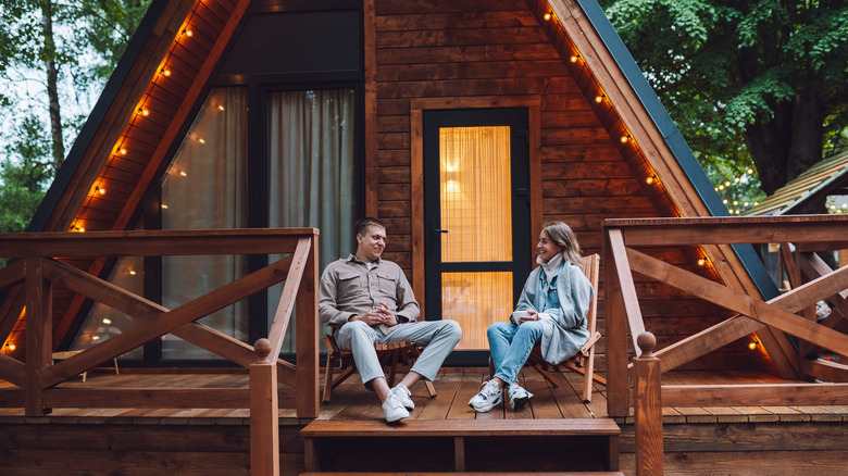 A man and woman happily sitting on the back porch of an A-frame wooden home nestled in the trees.