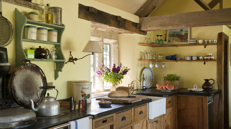 A rustic, country kitchen with pale yellow walls, a sage green shelf, and natural wood cabinets and beams. Fresh flowers, a loaf od bread, and watermelon slices are on the counter.