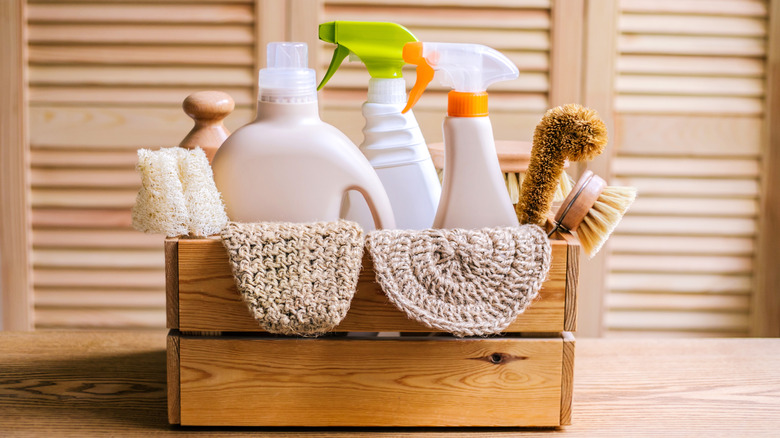 A box filled with neutral-colored cleaning supplies in front of a beige slated door.