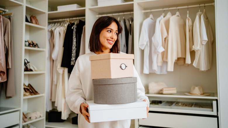 A smiling brunette woman standing in a beautifully organized closet, and holding 3 hat boxes.